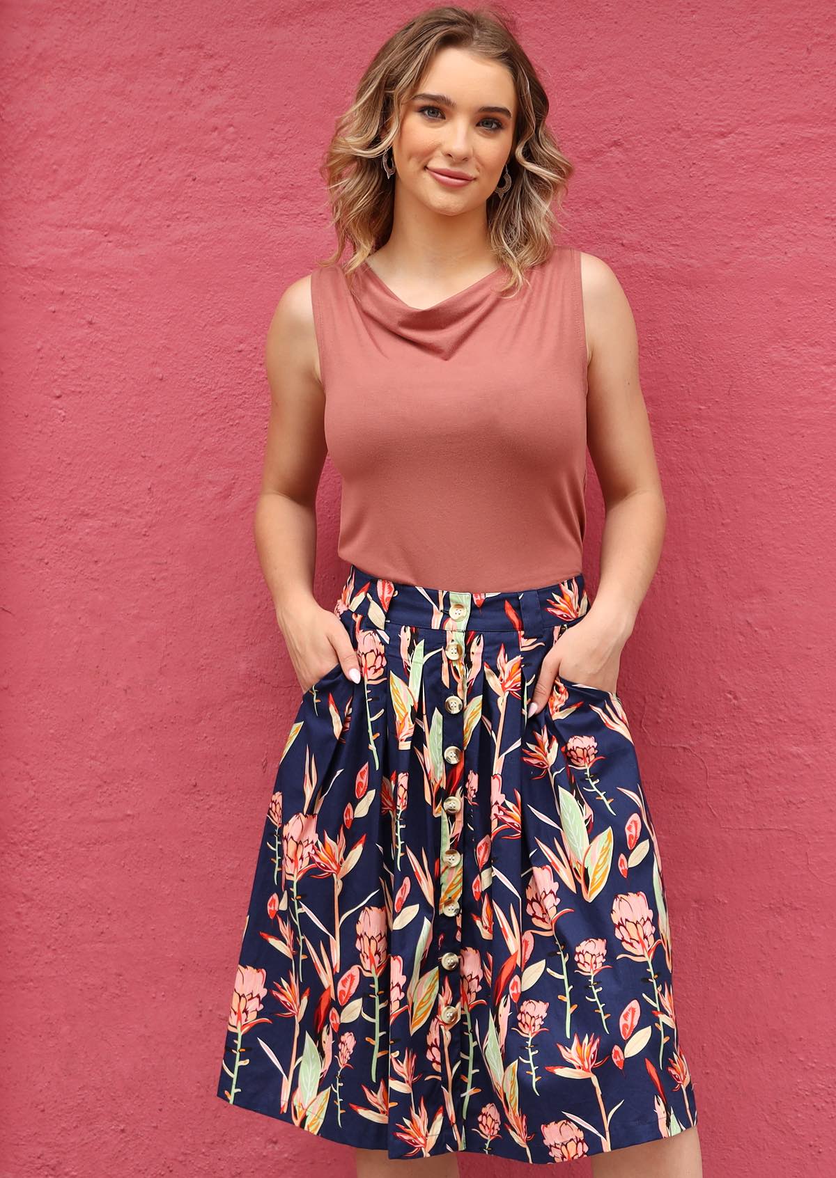 Woman wearing a pink sleeveless top and a navy floral button through knee length skirt against a pink wall, with hands in pockets