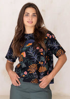 Woman in floral boxy style cotton top with short sleeves, round neckline and hands in green pants pocket.