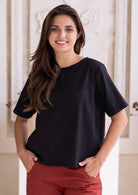 Woman wearing a black cotton seersucker t-shirt style top and rust pants, with hands in pockets