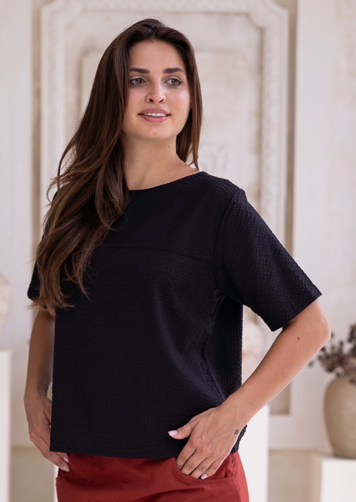 Woman wearing a t-shirt style black cotton seersucker top and rust pants, with hands in pockets