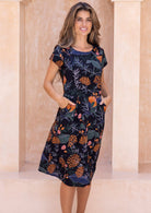 Model standing in a 100% cotton dress with hands in pockets of a beautiful botanical print dress with orange and pink florals.