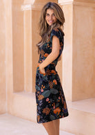 Model stands sideways with hands in pocket of a botanical print short sleeve sundress.