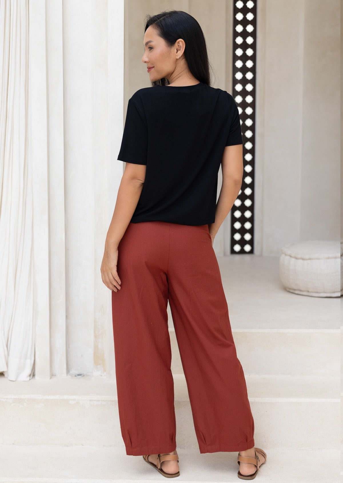 Back pose of model wearing burnt henna cotton pants with wide leg tapered fit, pleated ankles and pockets