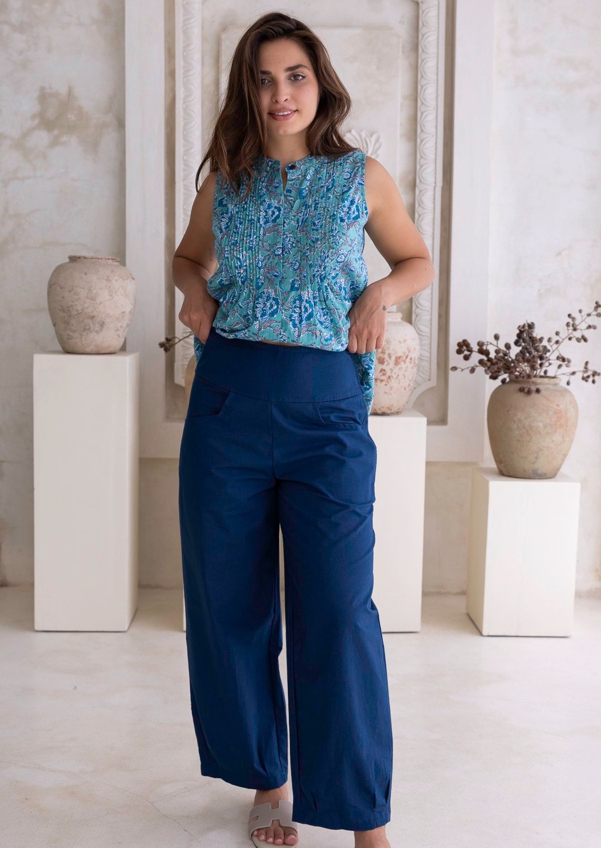 Woman wearing a deep sea blue relaxed cotton pants with aqua blue floral top in a minimalistic room with white walls and vases