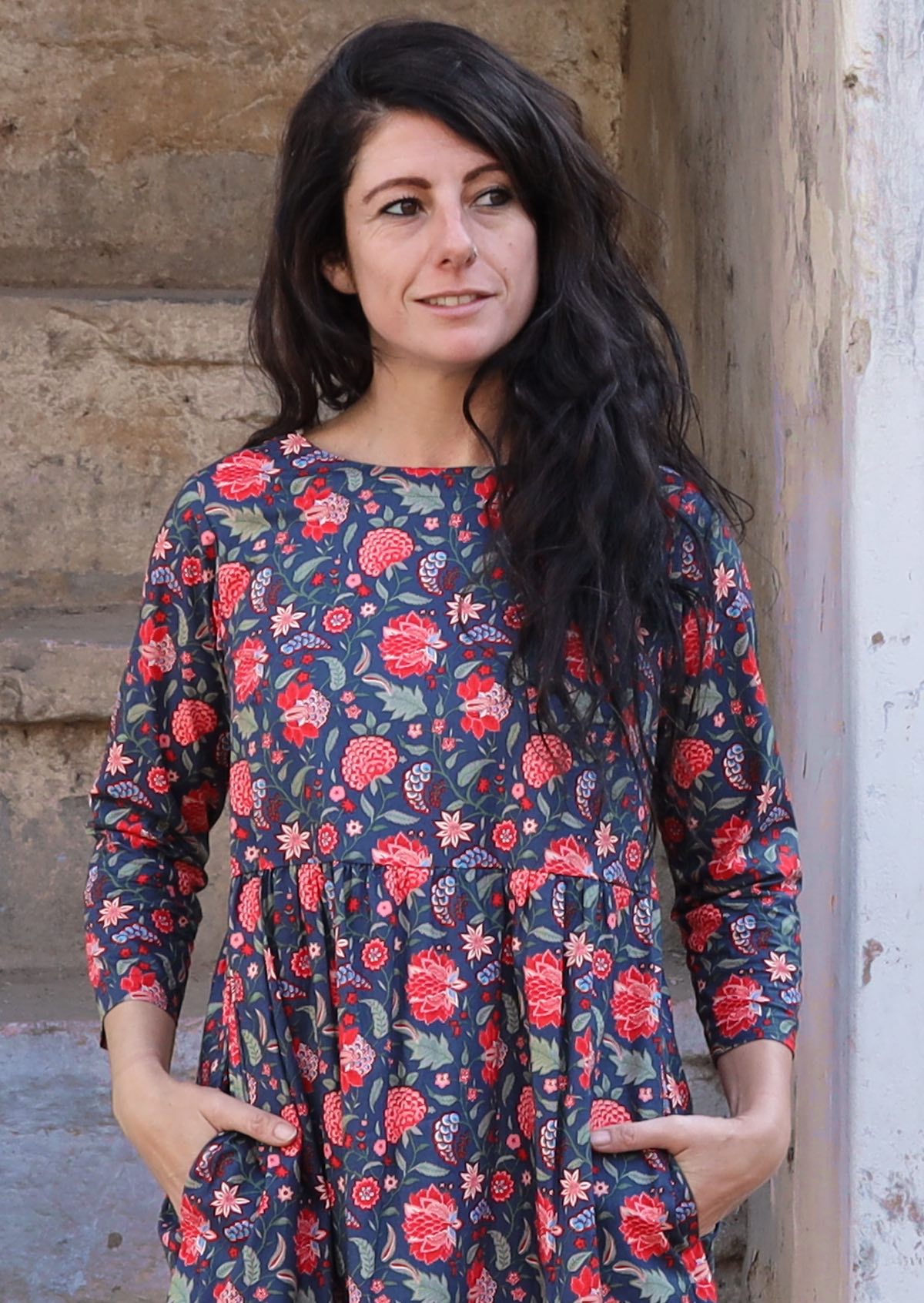 Close up of model with dark hair wearing a 100 percent cotton three tiered blue base and red floral print midi length dress with hands in pockets