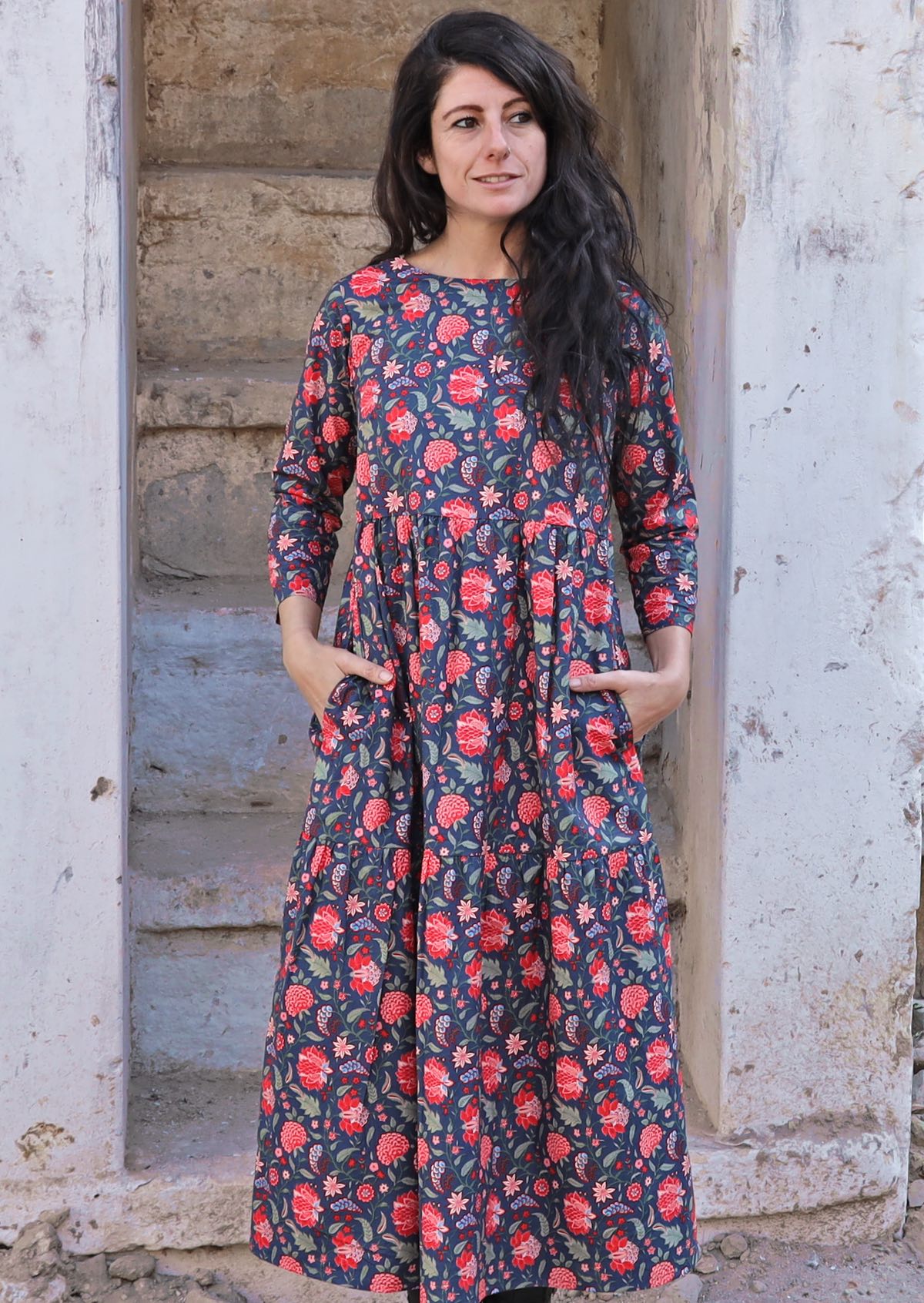 Model standing in front of cement stairs with dark hair wearing a 100 percent cotton three tiered midi length dress with blue base and red floral print with hands in pockets