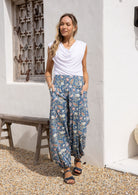 Woman wearing a white rayon top and a dusty blue floral pants with hands in deep pockets.