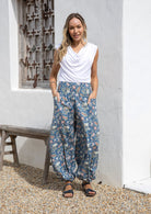 Woman wearing a white top and blue floral pants with wide elastic waist and ankles standing in front of a white wall with a wooden bench.