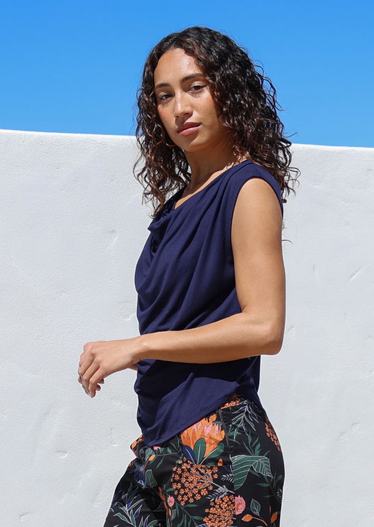 Woman wearing a navy sleeveless top and floral pants against a white wall with blue sky.