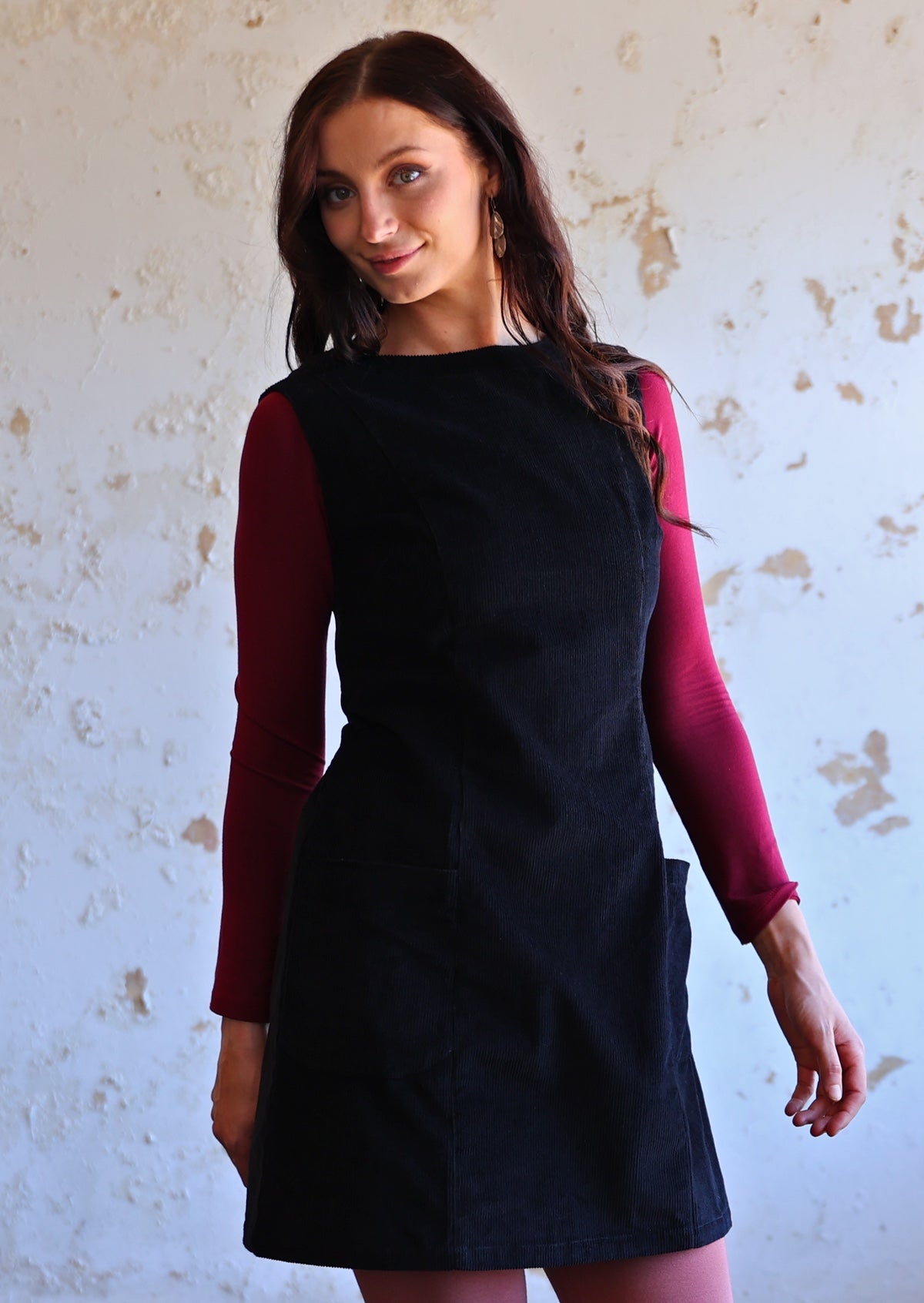 Woman wears cotton corduroy tunic in black with pockets, maroon long sleeve top underneath