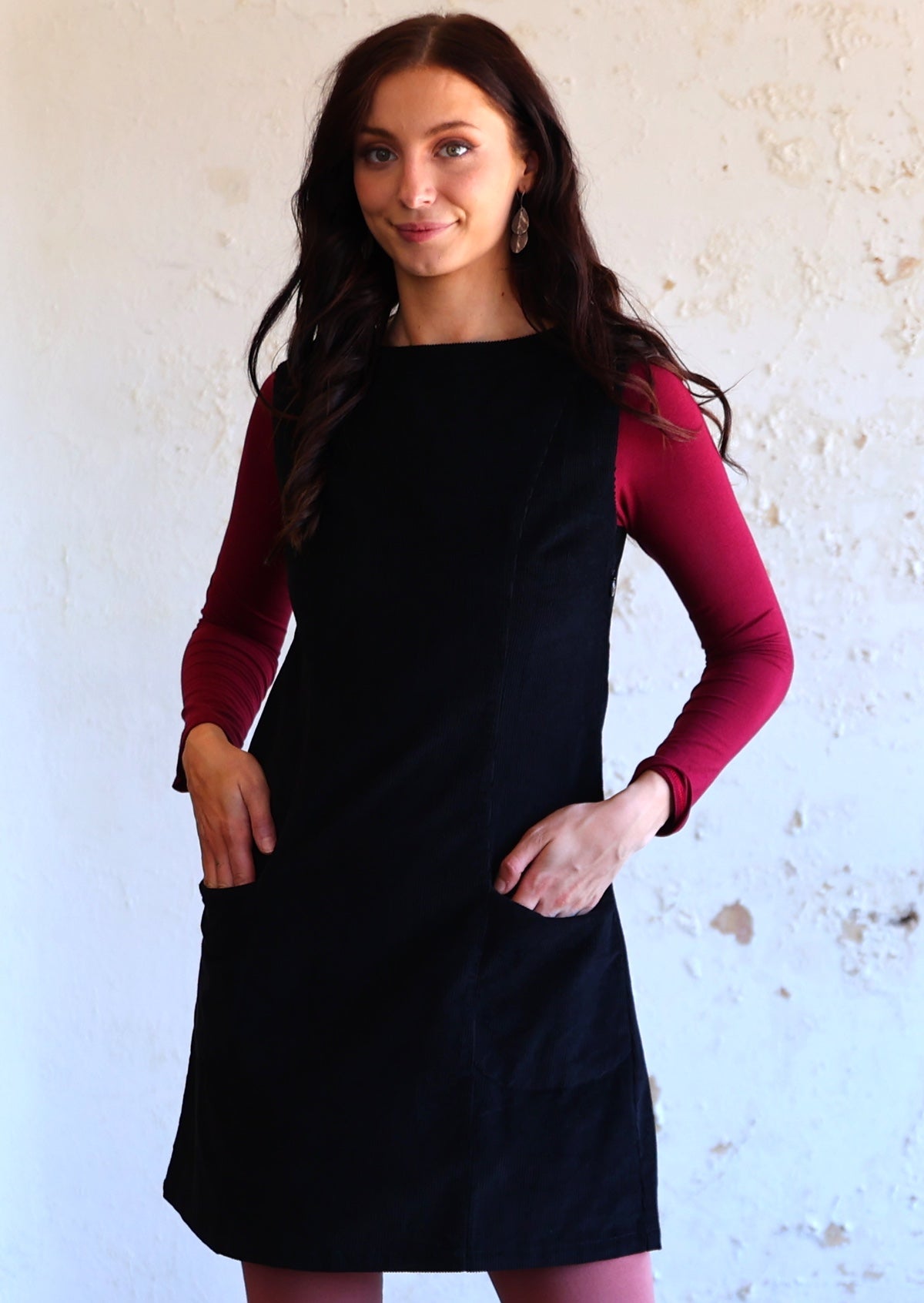 Woman wears cotton corduroy tunic in black with pockets, maroon long sleeve top underneath, hands in pockets