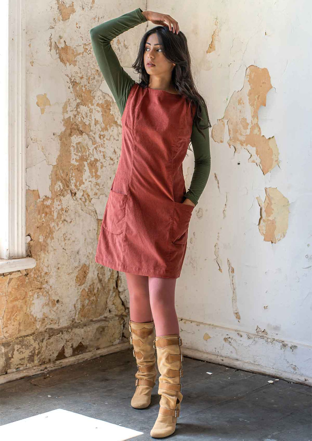 Woman wears burnt henna corduroy sleeveless above knee tunic with high round neckline, with her hand in pocket