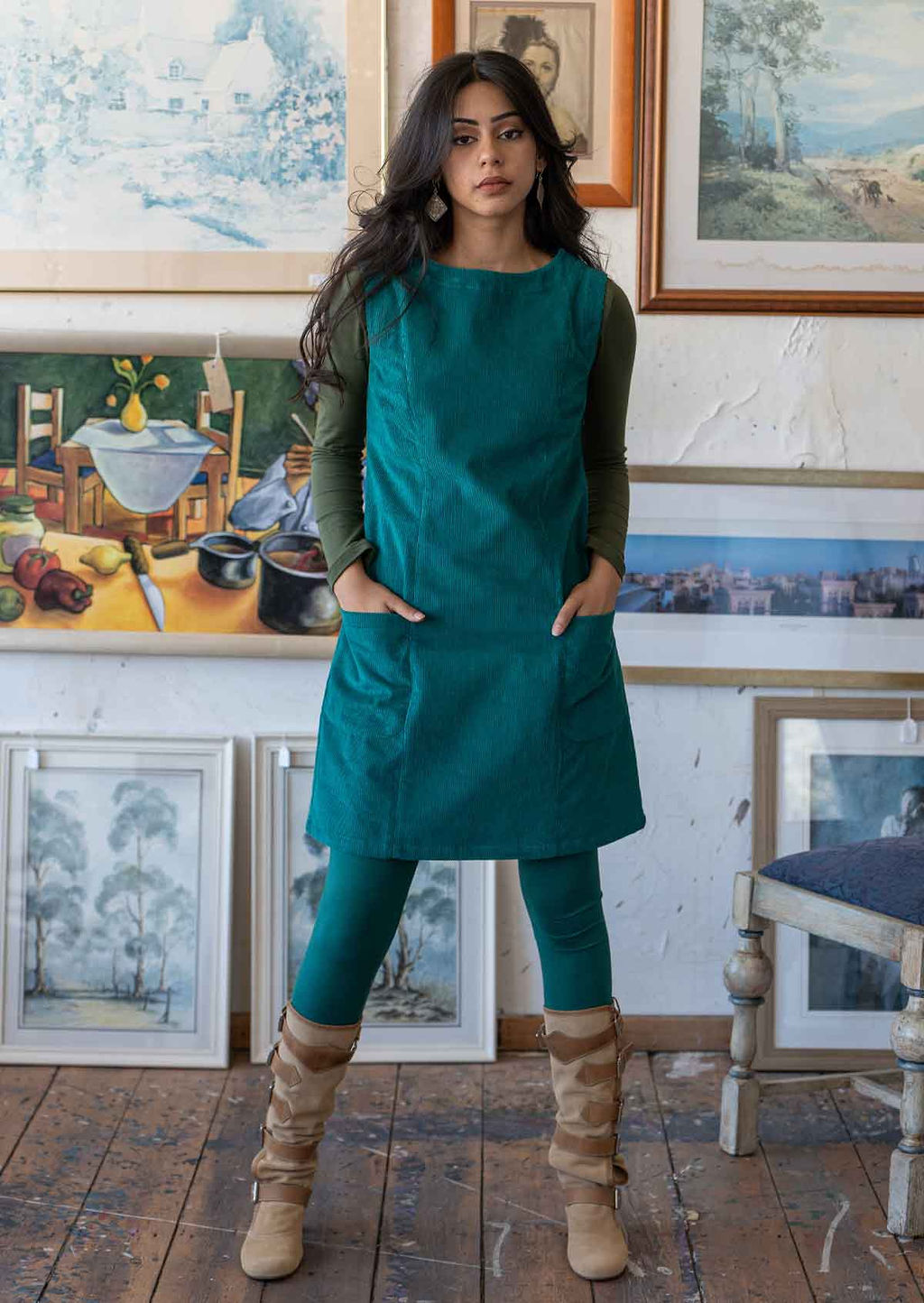 Model wears teal coloured corduroy sleeveless tunic with hands in pockets with an olive long sleeve top underneath, leggings and boots