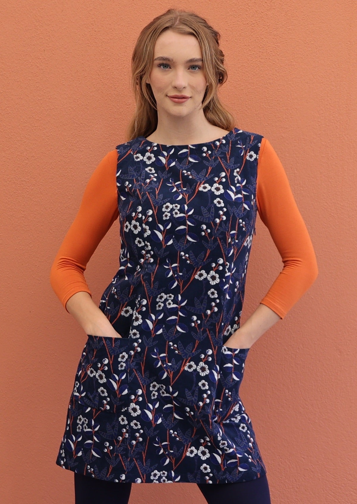 Woman wearing a sleeveless above knee length cotton corduroy tunic in orange, white and blue floral print on navy blue base.