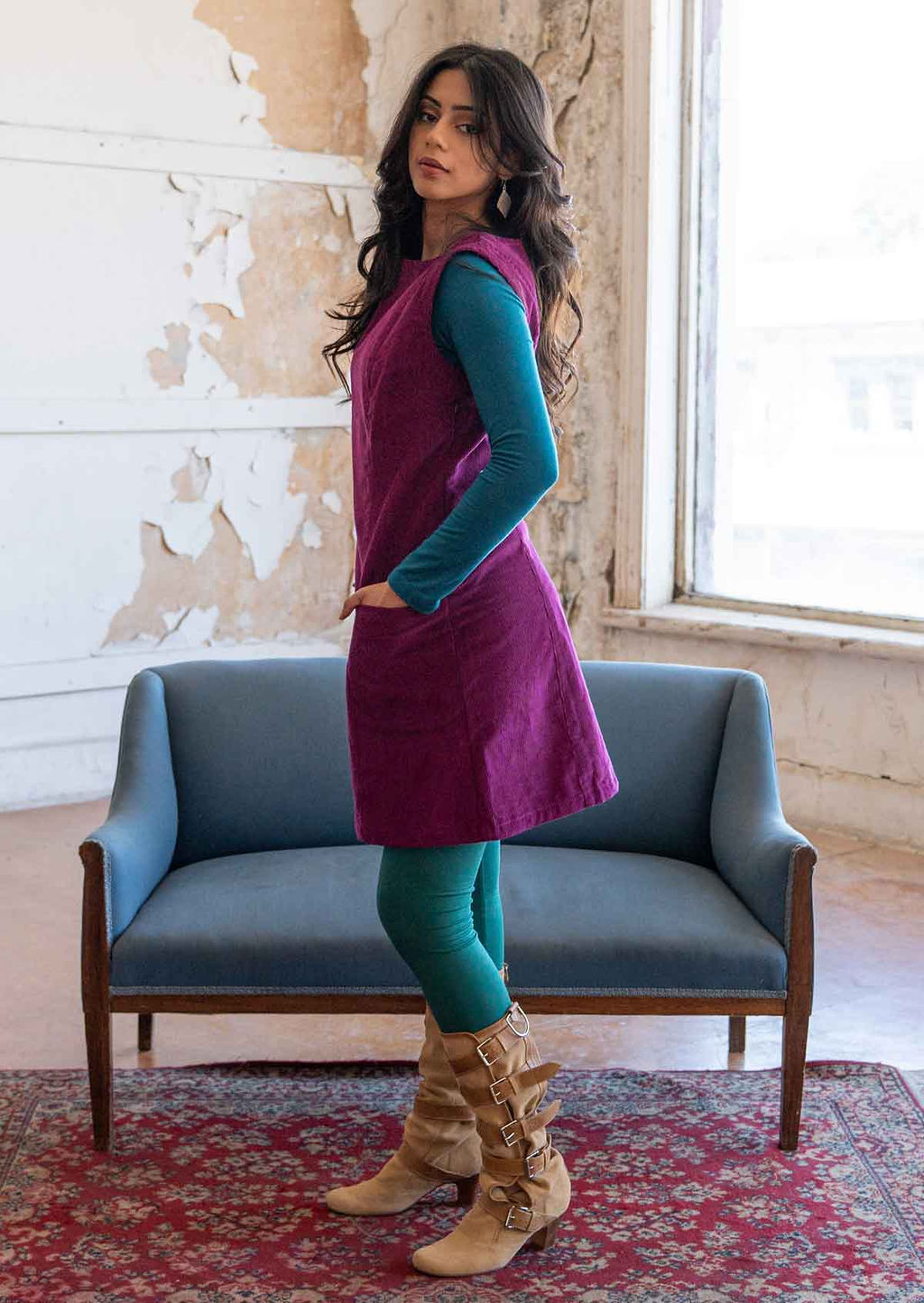 Model wears corduroy sleeveless tunic with hands in pockets,  she has a teal top underneath and jade leggings