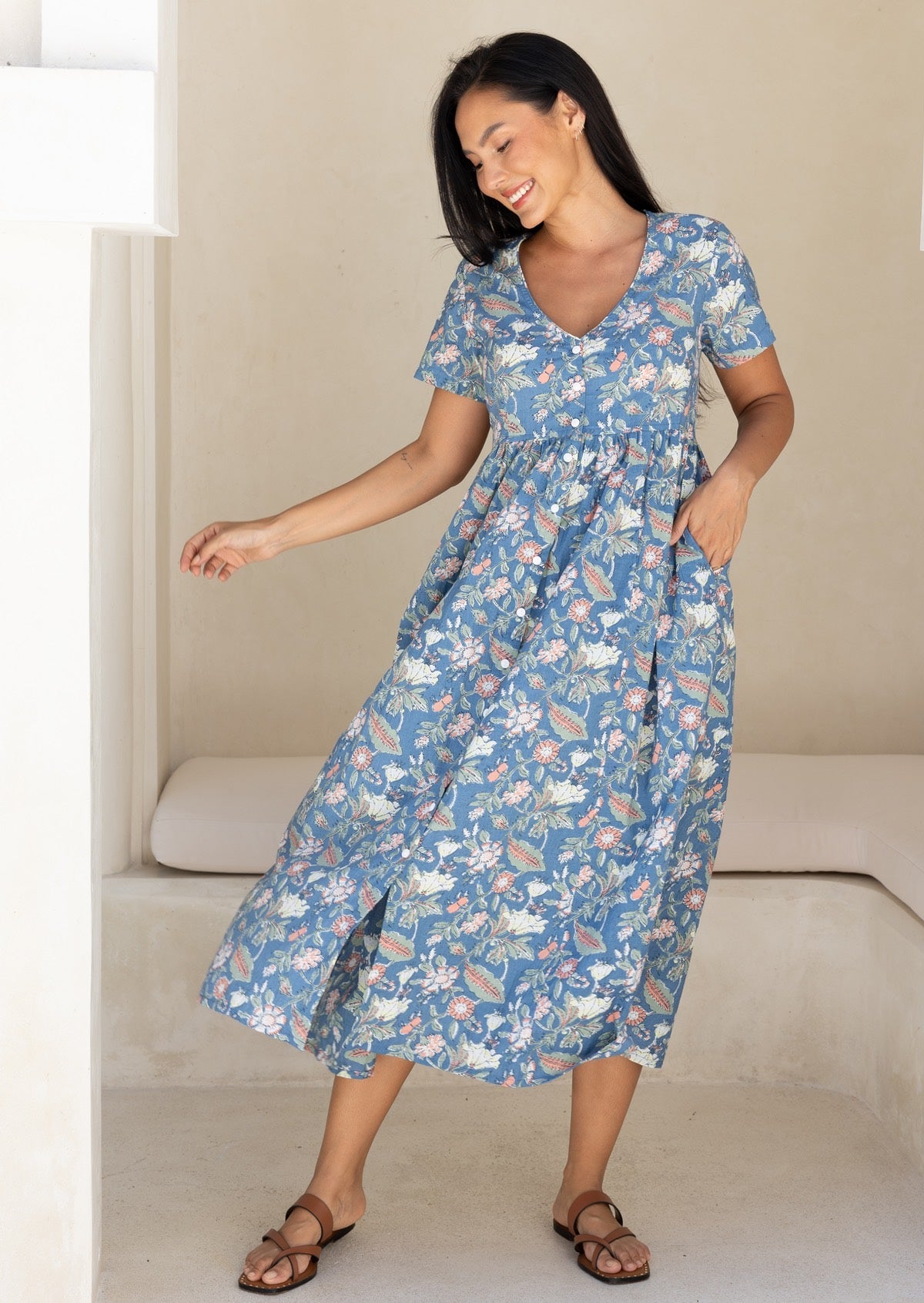 Woman wearing a blue based floral cotton midi length dress, with hand in pocket, swishing to one side