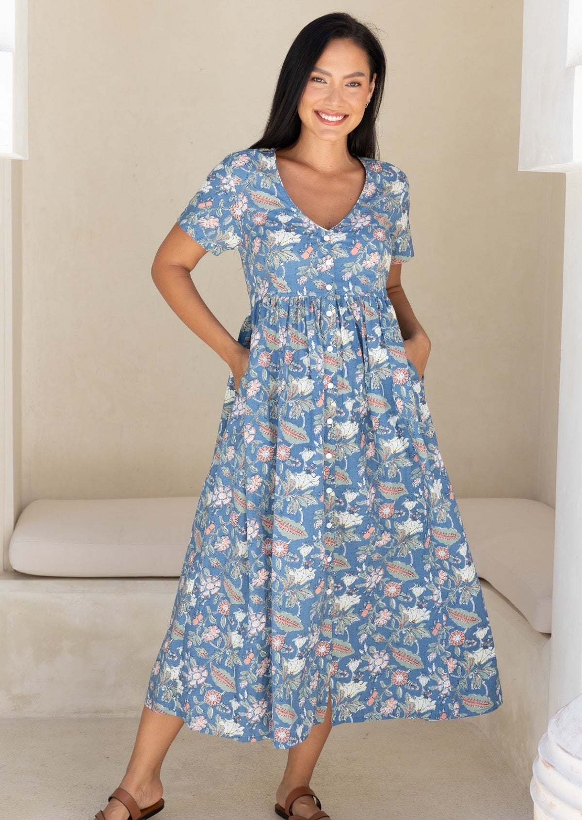 Woman wearing a blue based floral button through dress with a V-neckline and short sleeves, with hands in pockets