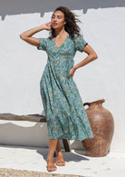 Woman wearing floral cotton midlength dress with short sleeves, shoestring tie and button front standing in front of white wall and wooden bench