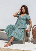 Woman sitting on a wooden bench in sage green floral cotton dress with button front, short sleeves, V neckline and pockets