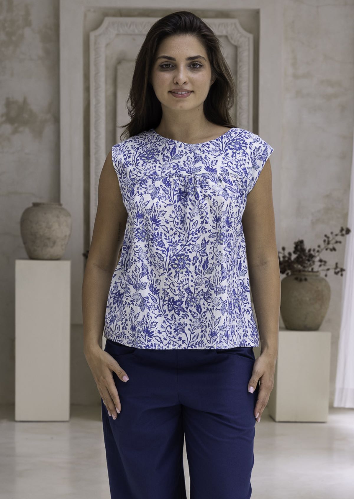 Woman wearing a white based blue floral cotton top with dark pants in an indoor setting.
