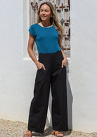 Woman wearing a blue top and black cotton wide legged pants standing against a white wall with hands in pockets