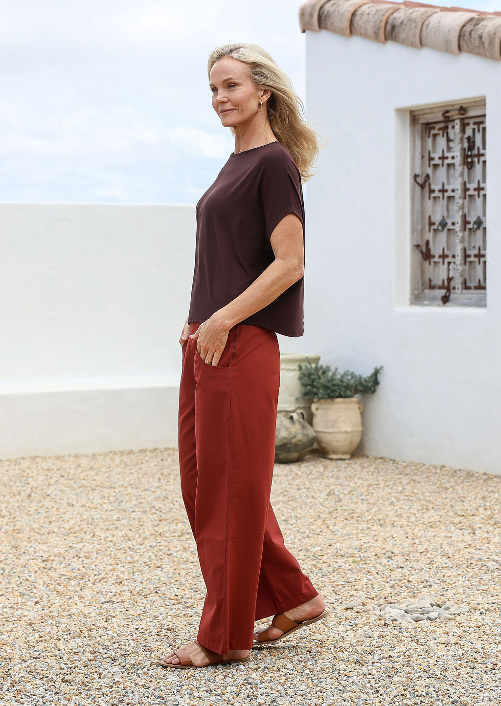 Woman wearing a brown top and burnt henna wide leg 100% cotton pants standing in front of a white wall with a wooden window.