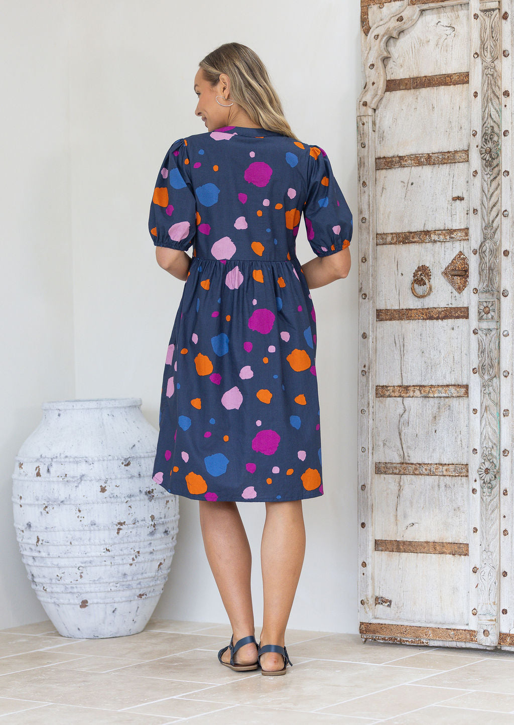 Back view of woman wearing a blue knee length relaxed fit dress with colourful polka dots standing in a white room
