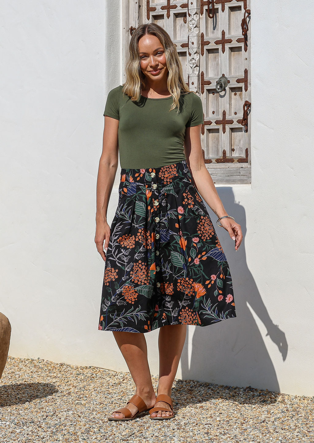 Woman wearing a green top and black A-line, button through cotton skirt with box pleats, belt loops and pockets 