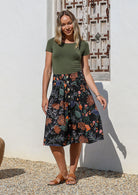Woman wearing a green top and black A-line, button through cotton skirt with box pleats, belt loops and pockets 