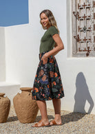 Model poses with hands in pockets wearing a knee length black cotton skirt featuring a lovely botanical print with belt loops, box pleats and elastic back waist