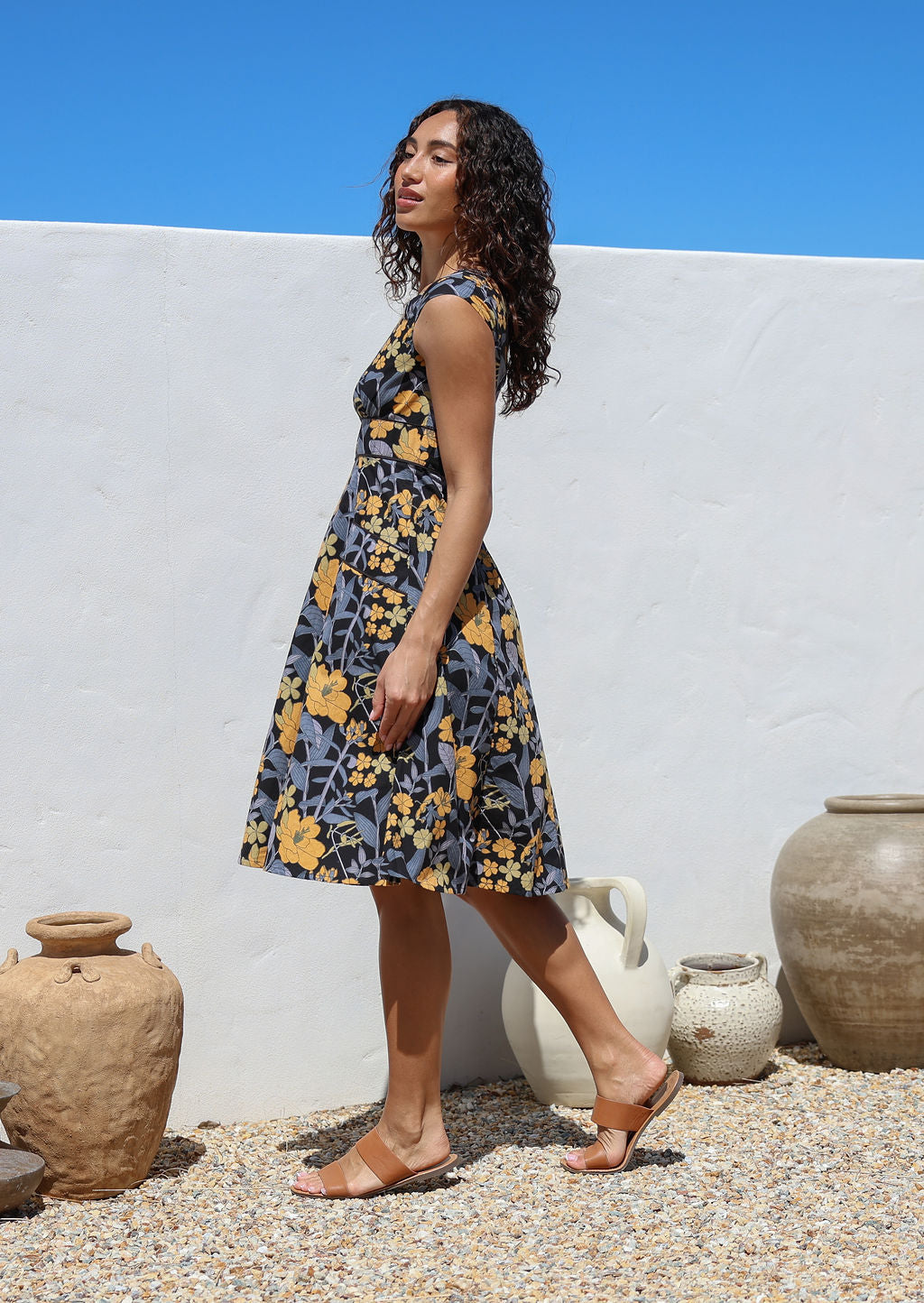 Model stands sideways infront of white wall wearing a cotton knee length retro floral print dress with cap sleeves, pockets and fuller A line skirt