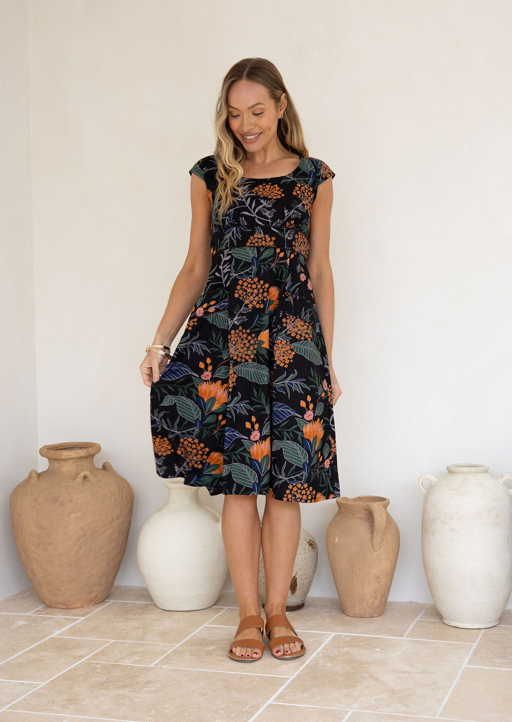 Model wearing a botanical print dress with scoop neck, cap sleeves and piped detailing at the waist and pockets.