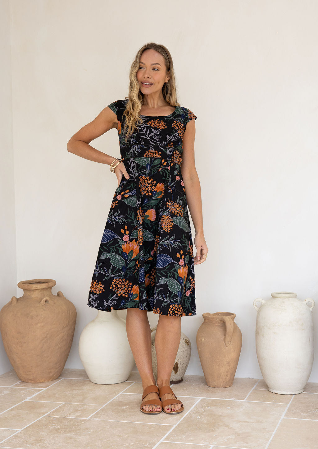 Model standing in front of white wall wearing a retro style 100 percent cotton dress with blue, green and grey leaves and pink, orange florals.
