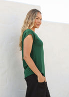 Woman stands sideways wearing a 100% rayon forest green shell t-shirt and black pants standing against a white wall