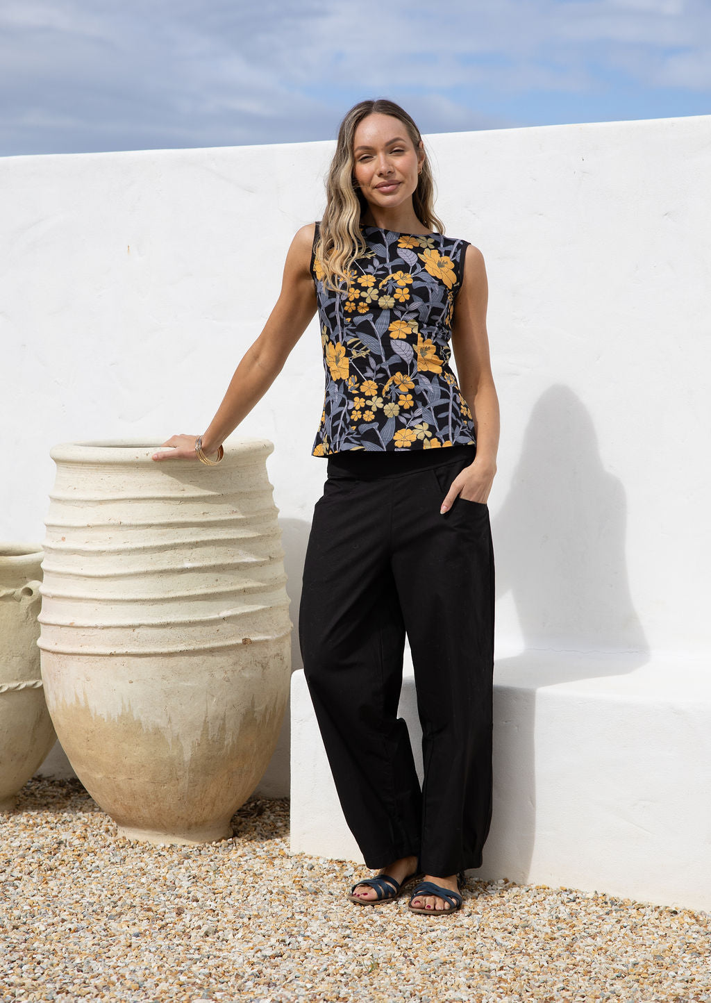Woman in floral sleeveless, tailored top and black pants standing against a white wall with large beige pot.