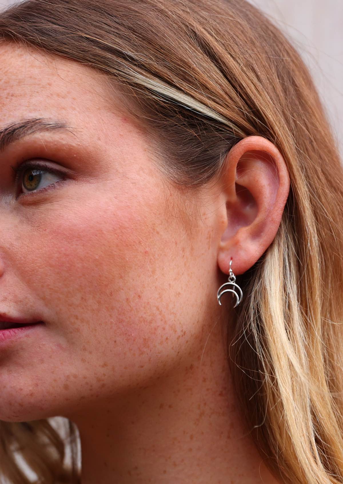 Woman wears silver linear crescent moon earrings, suspended from a wire hook