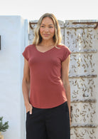 Woman with one hand in her pocket wearing a 100% rayon terracotta V-neck t-shirt and black pants standing against a textured wall.