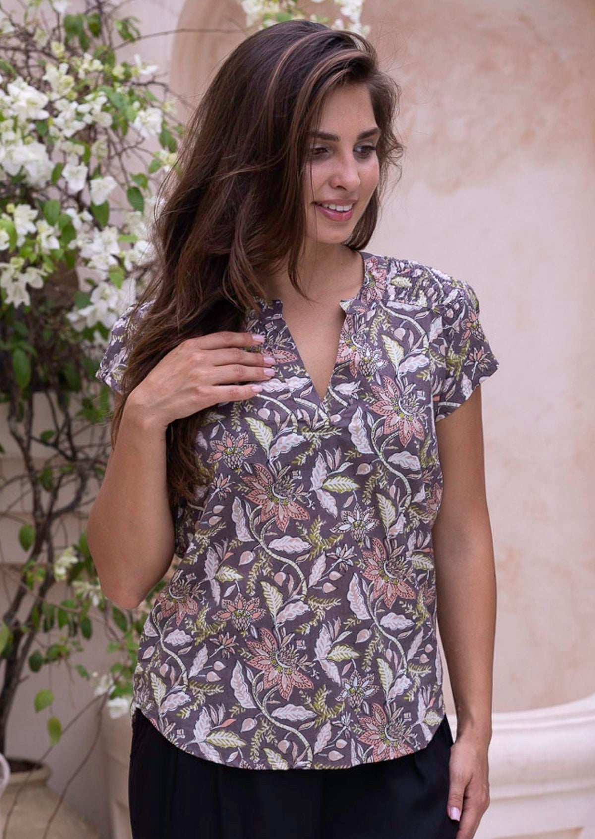 Woman wearing a light brown cotton blouse and black pants standing in an outdoor setting with decorative pots and plants.