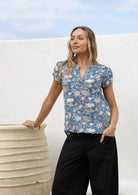 Woman posing in a dusty blue floral 100% cotton blouse with black cotton pants, standing against a white wall.
