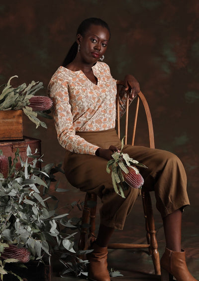 Model sitting down wearing tan cotton pants and cream floral blouse