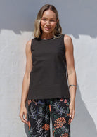 Woman wearing a relaxed fit black sleeveless cotton top with high round neckline, bust darts and trim detailing at armholes and neckline