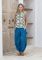 Woman wearing a colourful sleeveless top and blue pants standing in front of wooden doors.