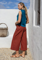 Woman in teal sleeveless top and rust wide leg, 3/4 pants standing against a white wall with a blue sky.