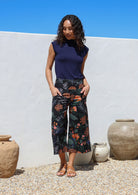 Woman standing with hands in pockets wearing a high waisted wide leg capri style pants featuring beautiful botanical print with pink and orange florals