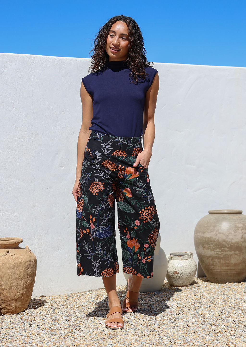 Woman stands with one hand in pocket wearing a black based botanical print wide leg, 3/4 length cotton pants with side zipper and pockets