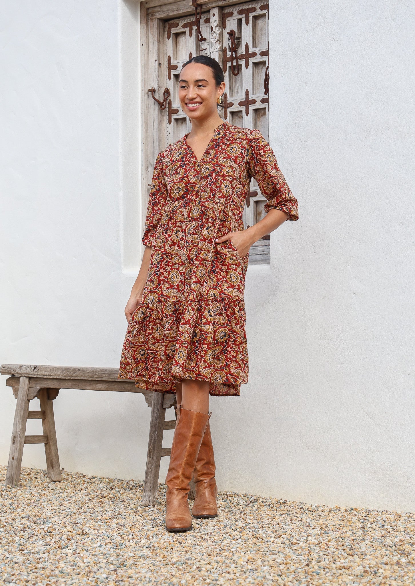 Woman wears cotton tiered dress in bright orangey red based floral print, with 3/4 sleeves, buttoned bodice and V-neckline, with hand in pocket