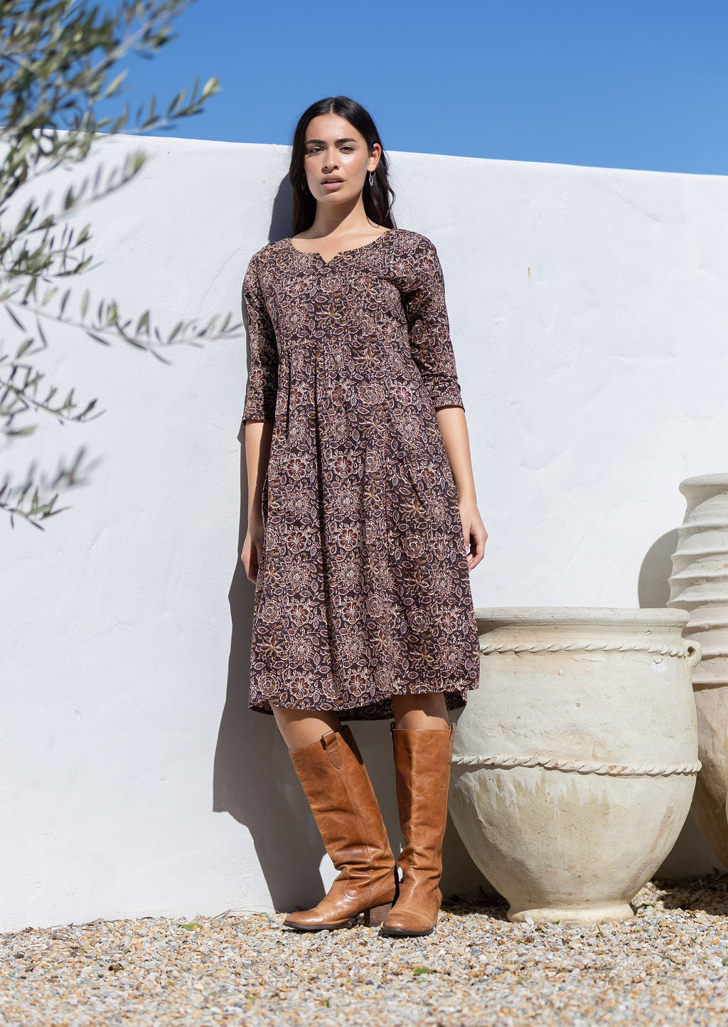Woman wearing a brown cotton patterned dress and brown knee-high boots standing against a white wall with an olive tree.