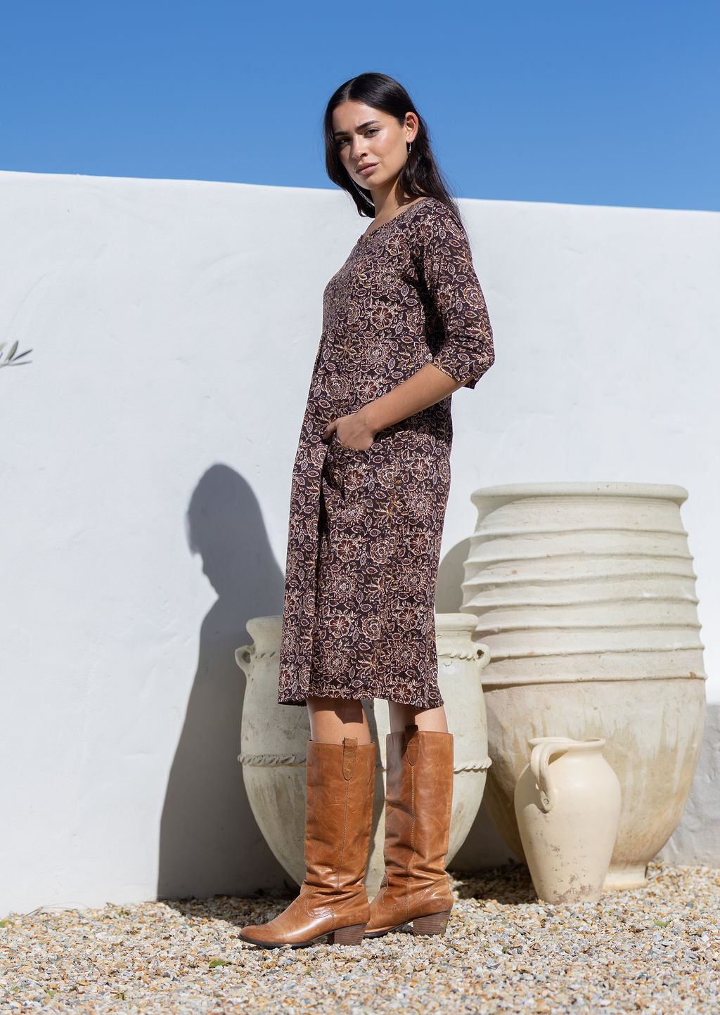 Woman in a brown floral cotton patterned dress and brown boots standing against a white wall with hands in pockets 