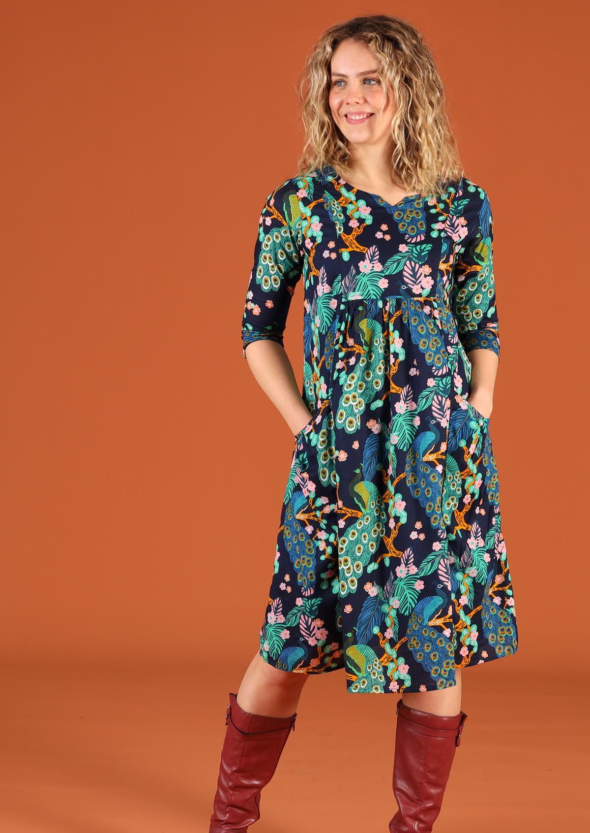 Woman models a 3/4 sleeved, knee length cotton dress in a peacock print with a dark blue base