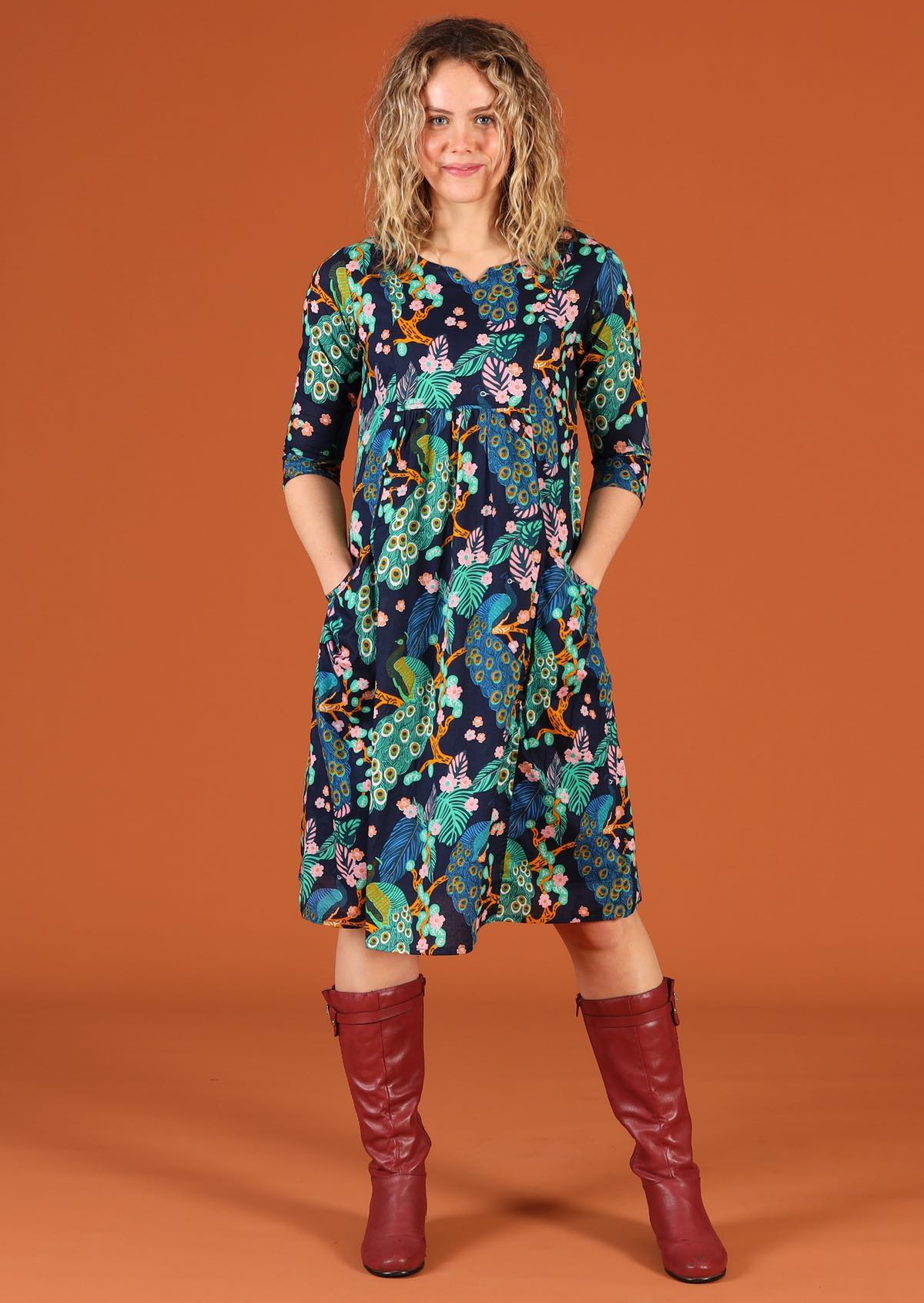 Woman models a knee length cotton dress with 3/4 sleeves in a peacock print, with hands in pockets